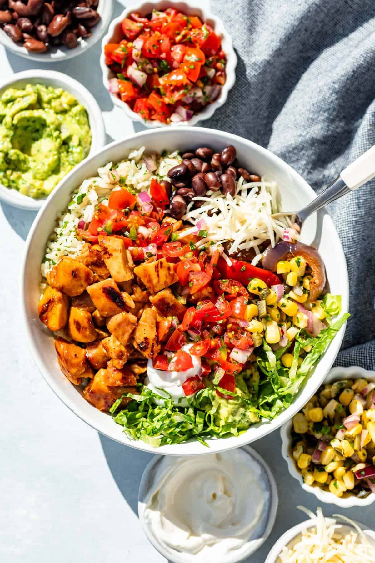 A white bowl filled with romaine, rice, Chipotle honey chicken, corn salsa, salsa, guacamole, cheese, and black beans with small bowls of salsa, guacamole and cheese around it.