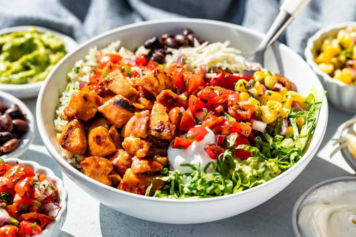 A chipotle honey chicken burrito bowl with chicken, rice, romaine, cheese, black beans, corn salsa, guacamole, salsa, and sour cream with extra bowls of toppings around it.
