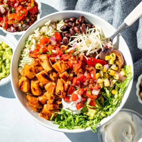 A chipotle honey chicken burrito bowl with chicken, rice, romaine, cheese, black beans, corn salsa, guacamole, salsa, and sour cream with extra bowls of toppings around it.