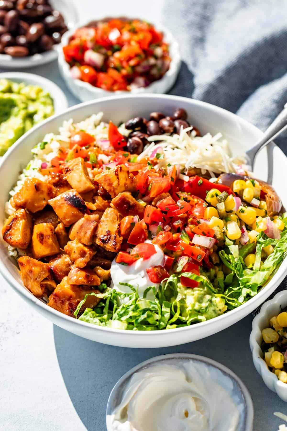 A Chipotle honey chicken burrito bowl with small bowls of Pico de Gallo, corn salsa, sour cream, and black beans around it.