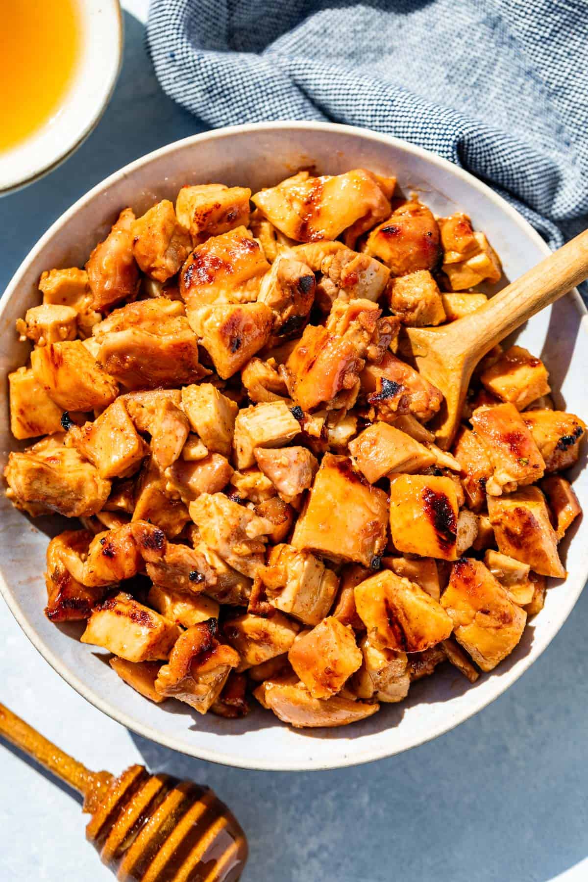 A bowl of chopped chipotle honey chicken with a wood spoon in it and a honey stick on the side.