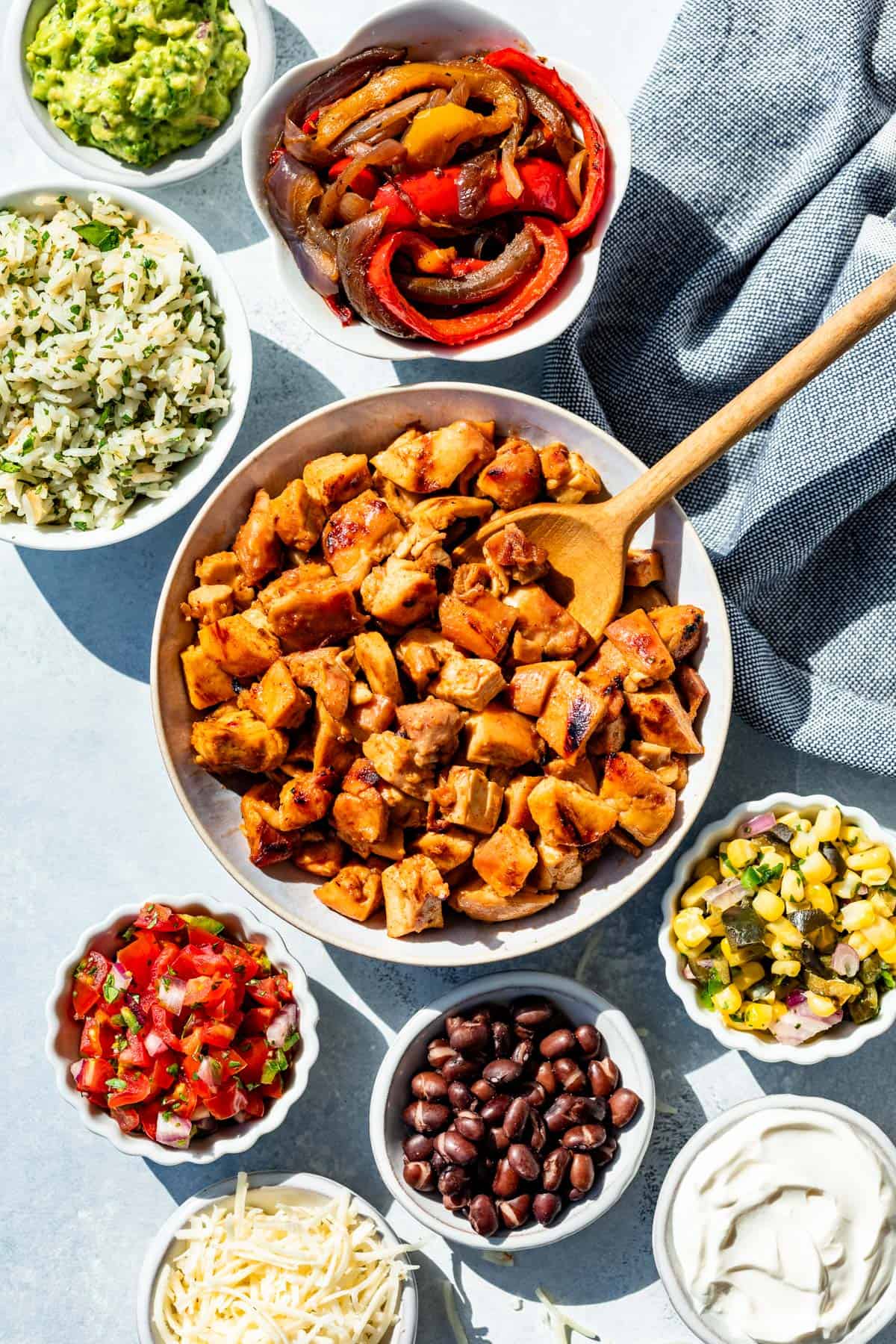 A bowl of Chipotle Honey Chicken on a blue background surrounded by bowls of rice, Fajita veggies, black beans, Pico de Gallo, Guacamole, Corn Salsa, Grated cheese, and sour cream.