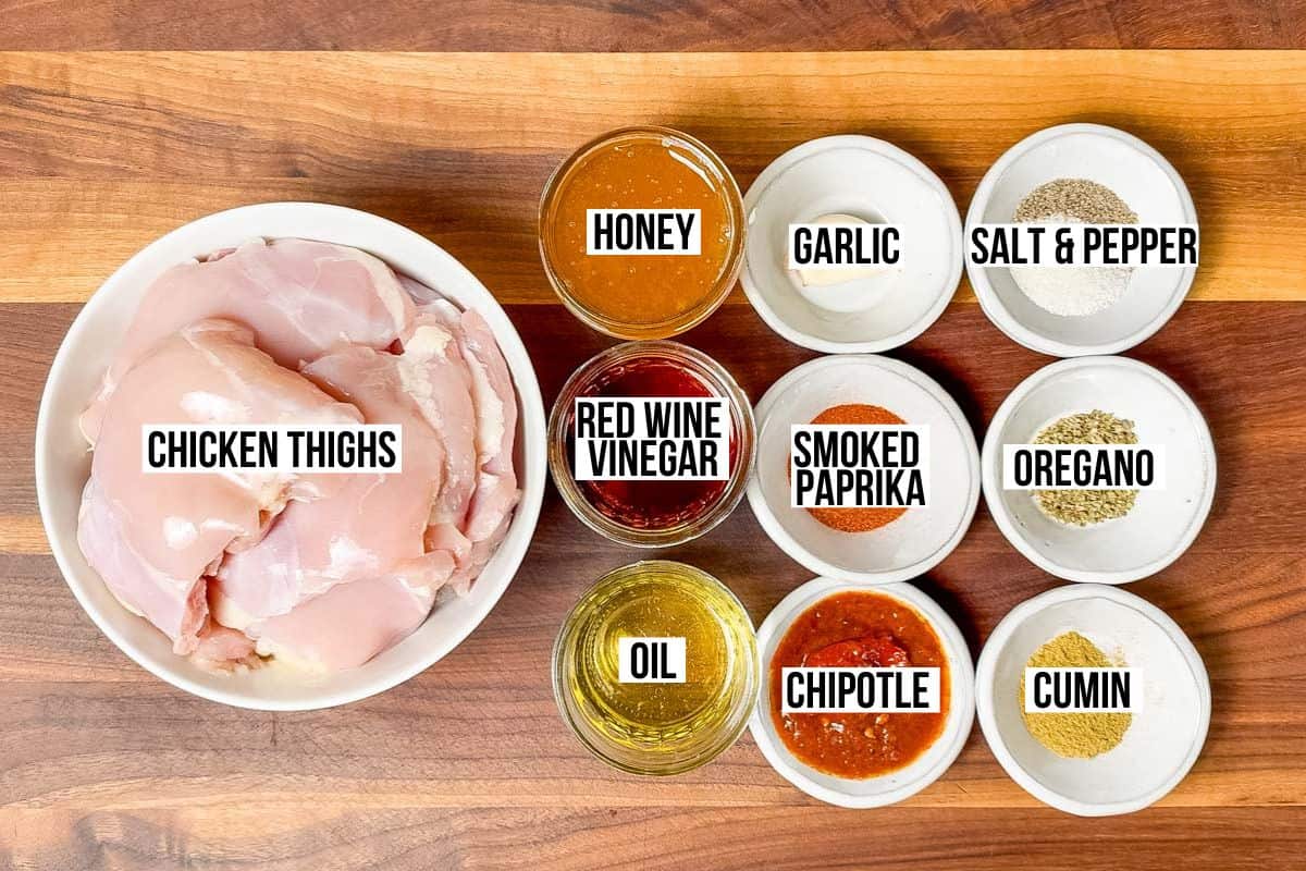 Bowls of chicken thighs, red wine vinegar, honey, oil, smoked paprika, oregano, garlic, and chipotle on a wood cutting board.
