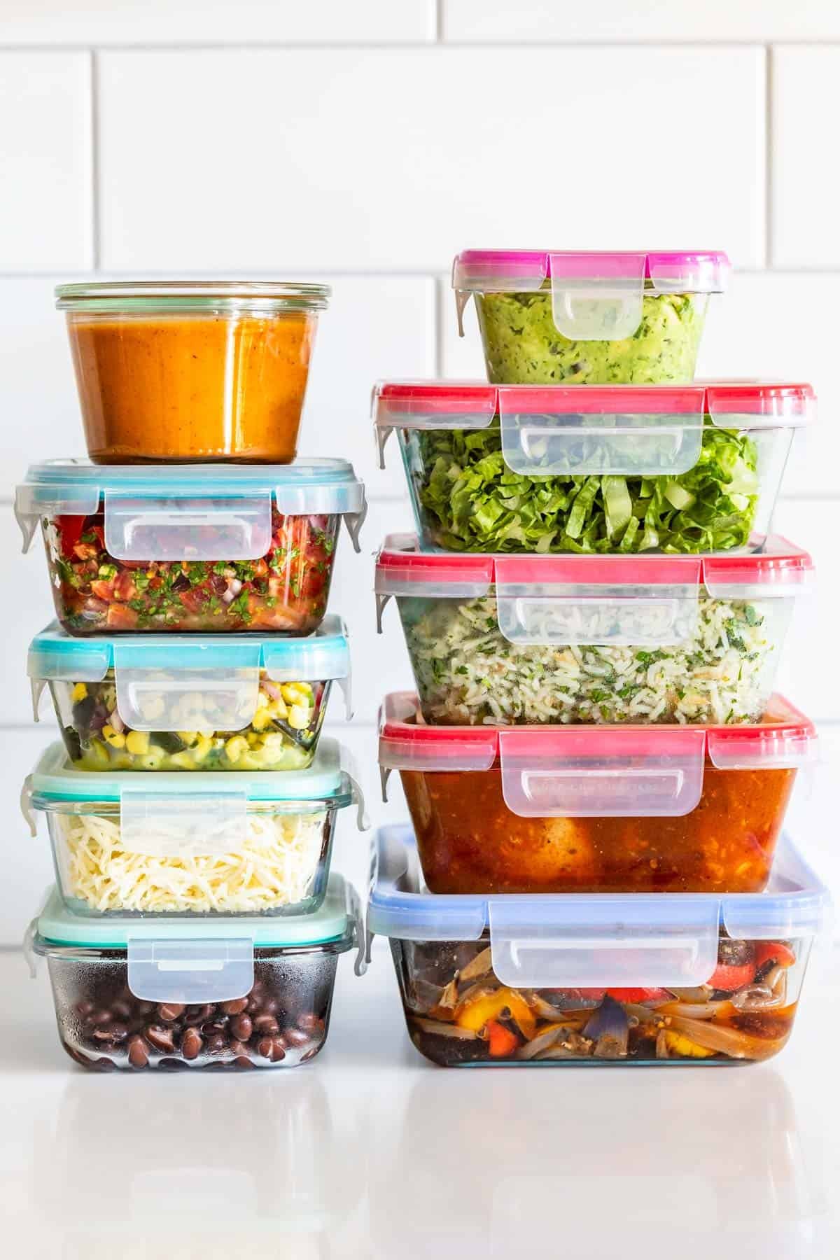 Glass containers of black beans, salsa, cheese, guacamole, lettuce, marinated chicken, fajita veggies, and rice on a white countertop.