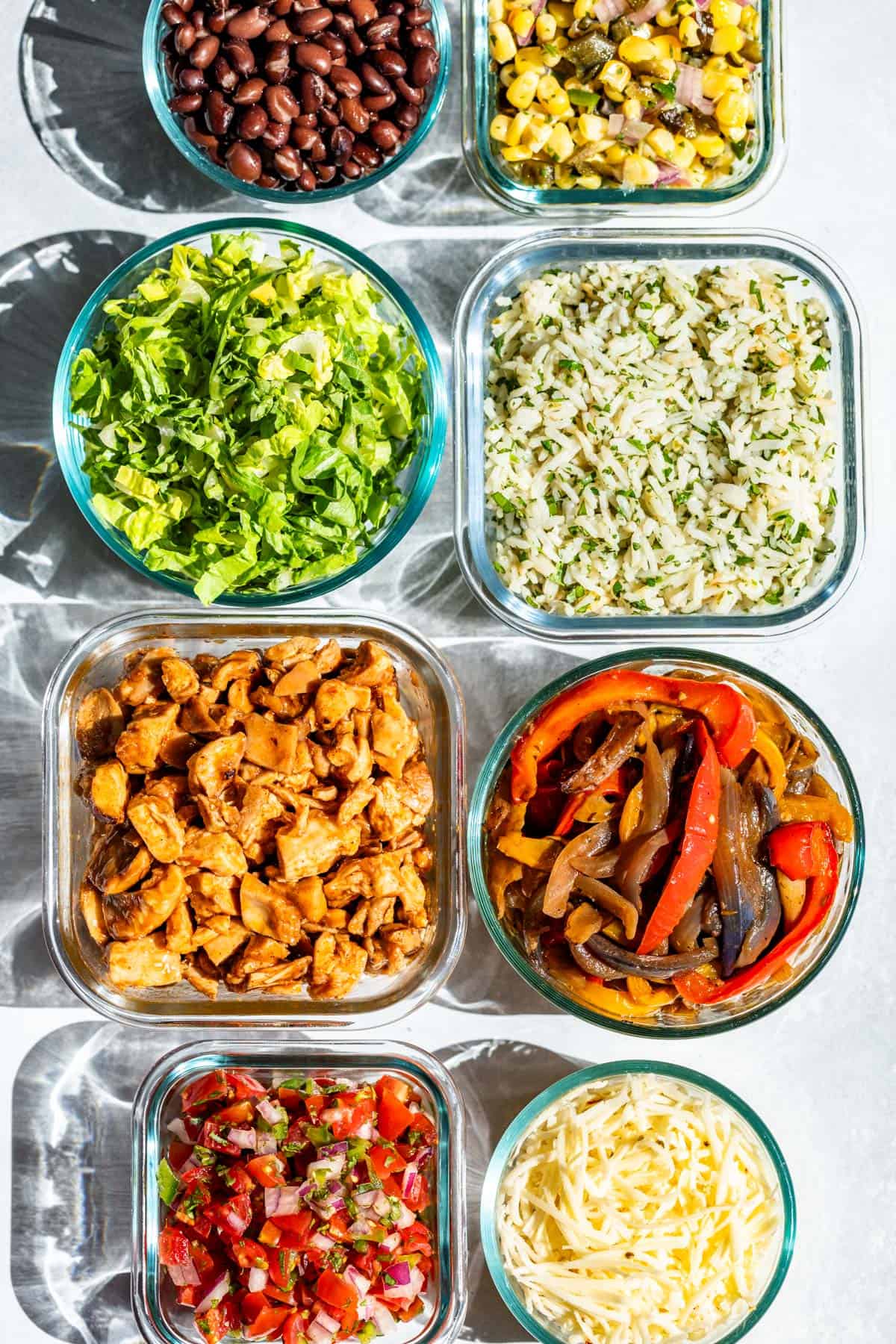 Glass bowls of black beans, Chipotle honey chicken, cilantro lime rice, Fajita veggies, Pico de Gallo, Chili Corn Salsa, grated cheese, and shredded romaine lettuce.