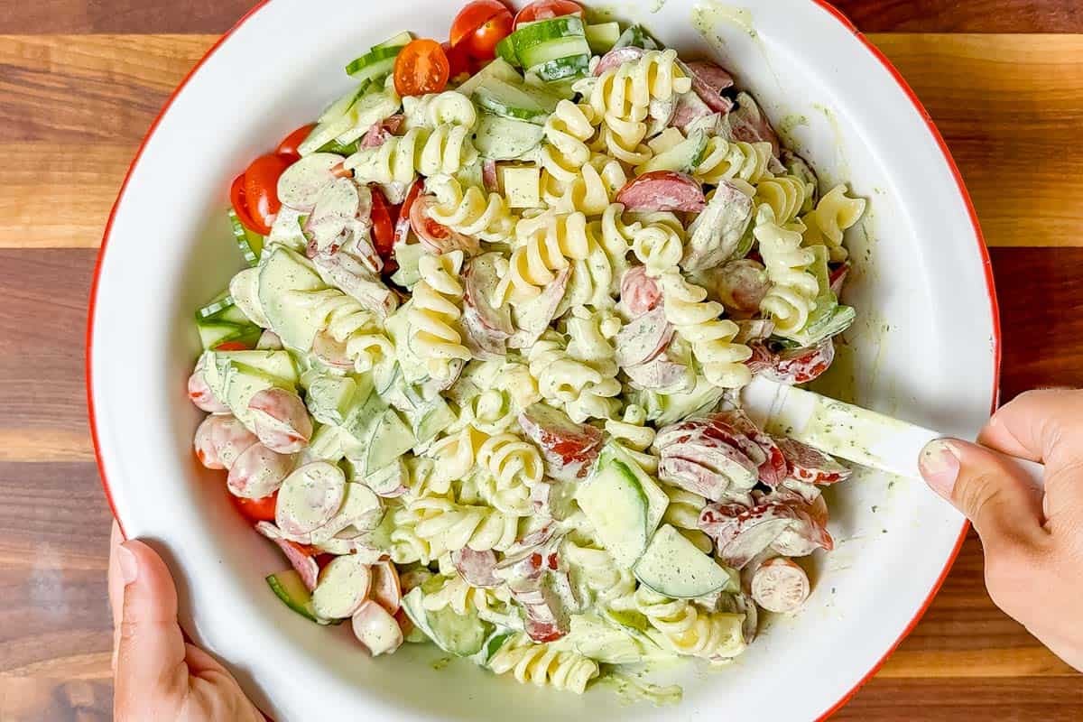 Mixing the pasta salad ingredients together with the creamy basil dressing in a red rimmed large white bowl.