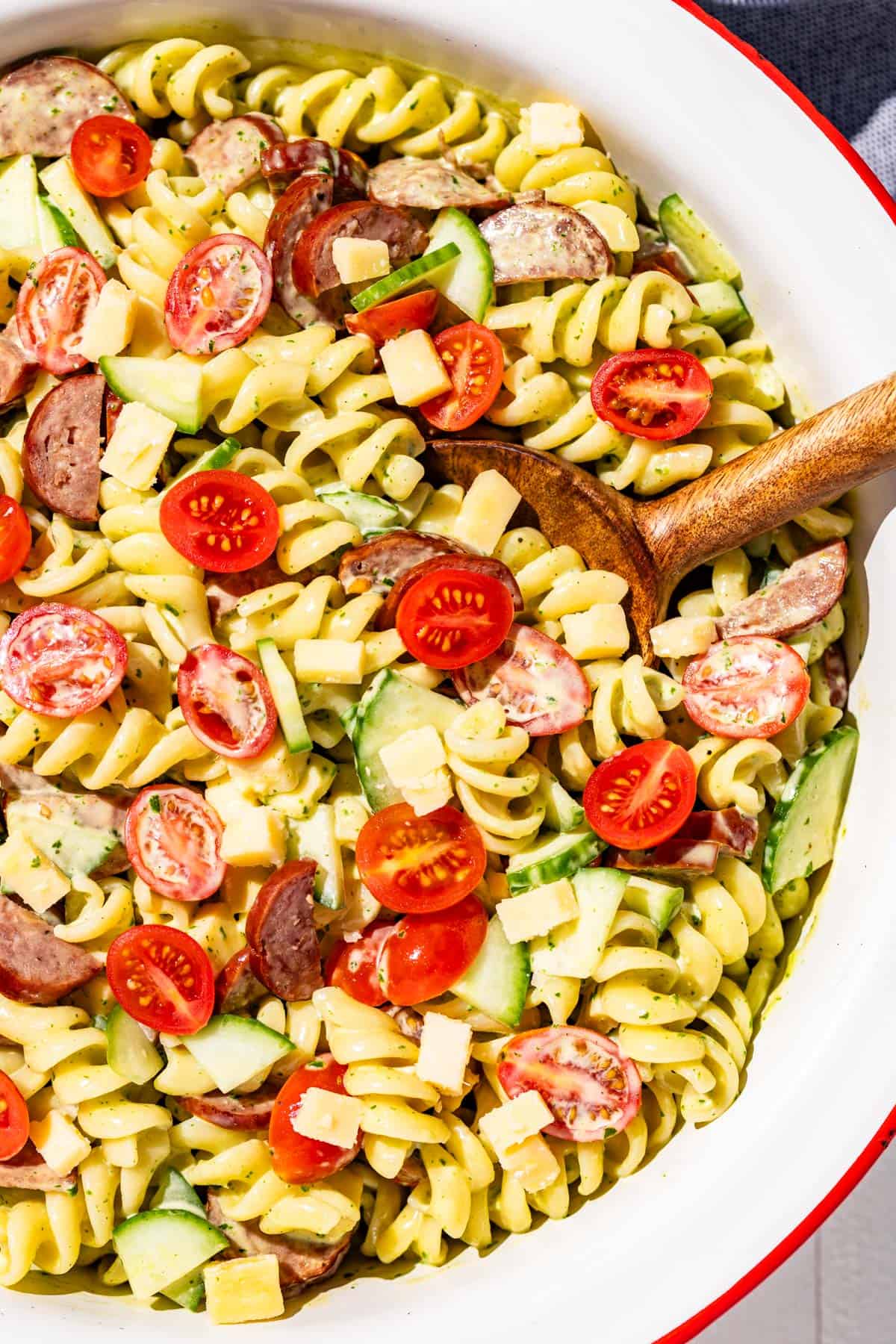 A large white bowl with a red rim filled with pasta salad with a wood spoon in it.