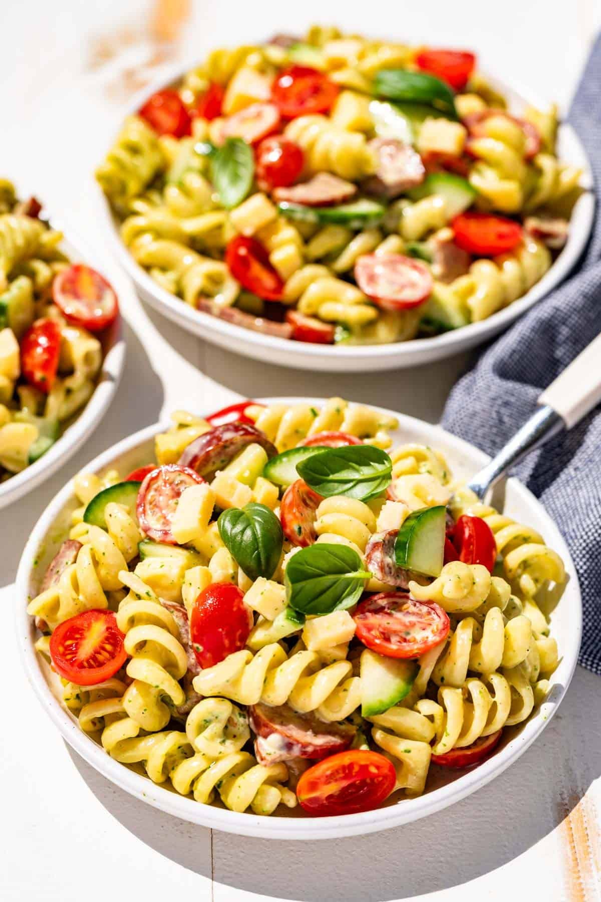 Three white plates filled with creamy pasta salad with a white handled fork in the pasta salad and a blue linen on the side.
