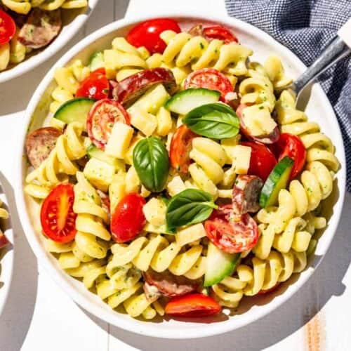Finished creamy pasta salad on three white plates topped with extra tomatoes and basil leaves with a white handled fork on the side.