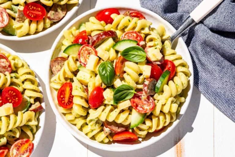 Finished creamy pasta salad on three white plates topped with extra tomatoes and basil leaves with a white handled fork on the side.