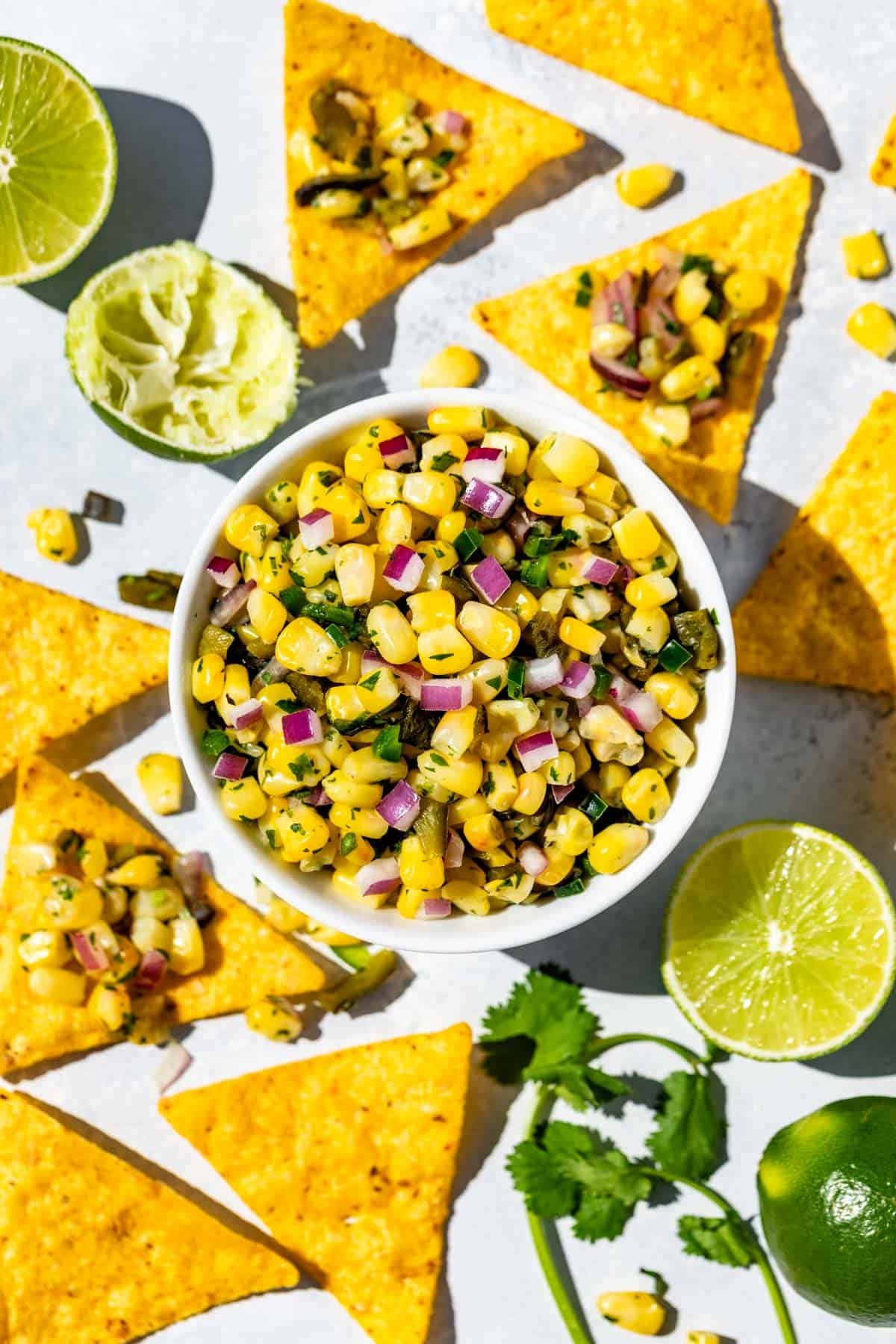 Roasted Chili Corn Salsa in a white bowl surrounded by corn chips, cilantro sprigs and limes.