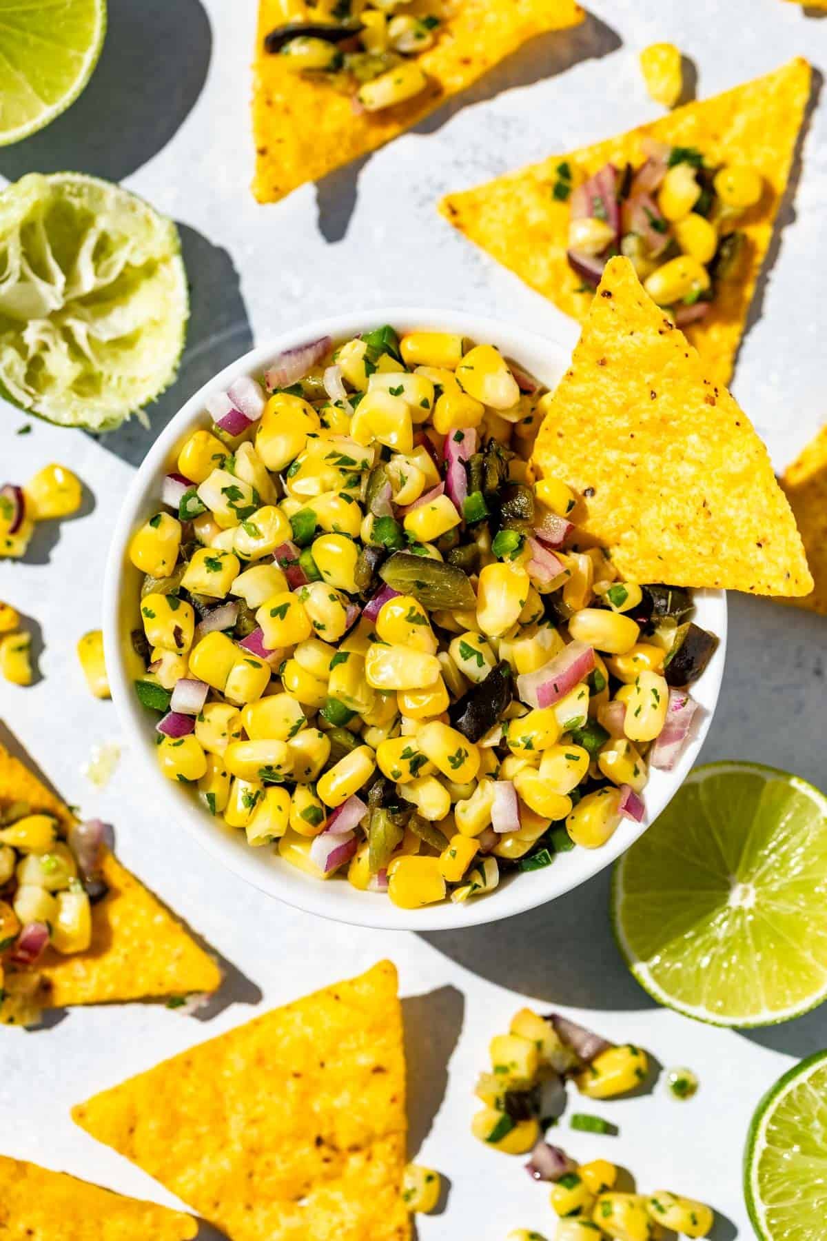 Roasted Chili Corn Salsa in a white bowl with a corn chip dipping into it surrounded by corn chips, limes, and cilantro.