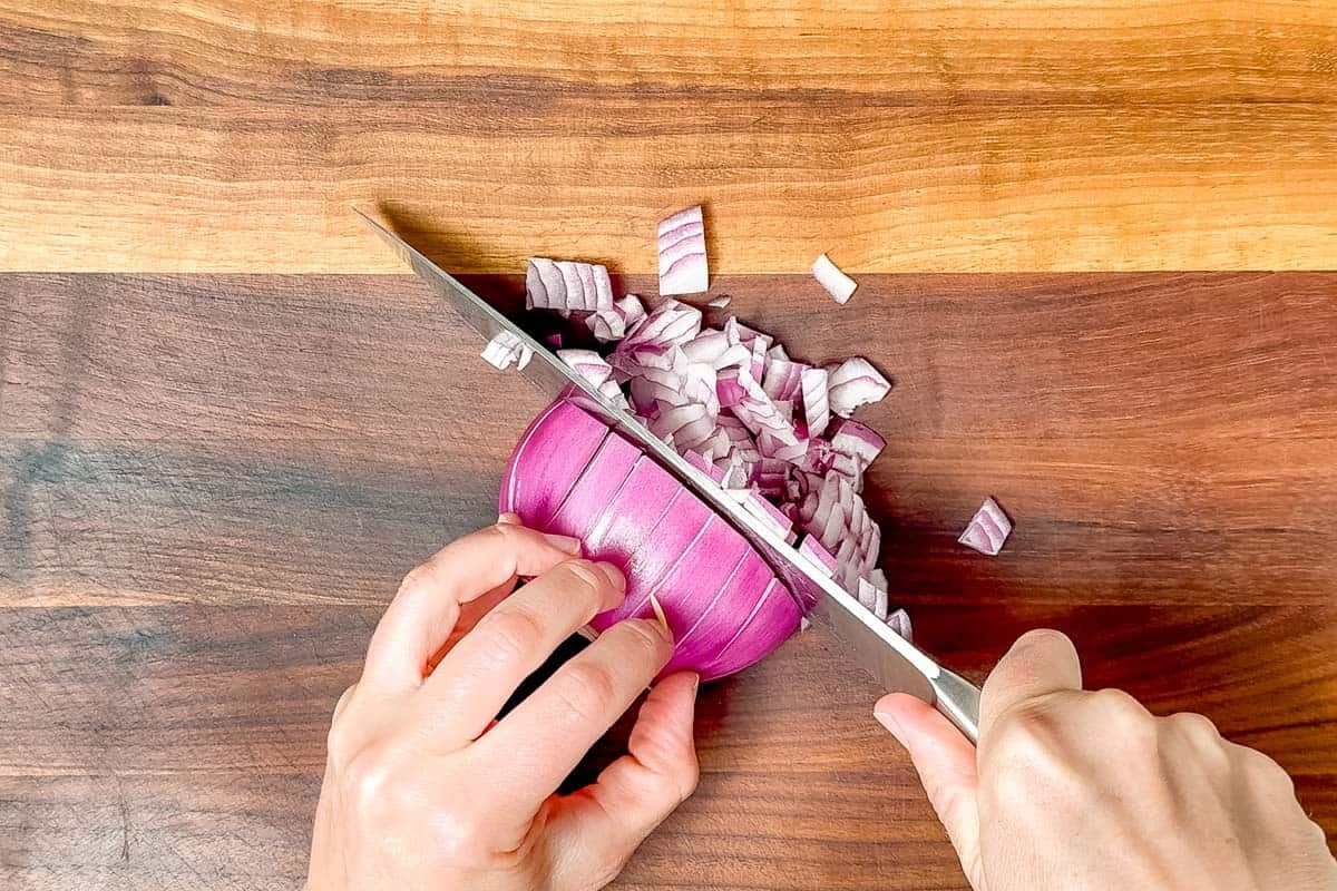 Finely dicing the red onion with a chef's knife on a wood cutting board.