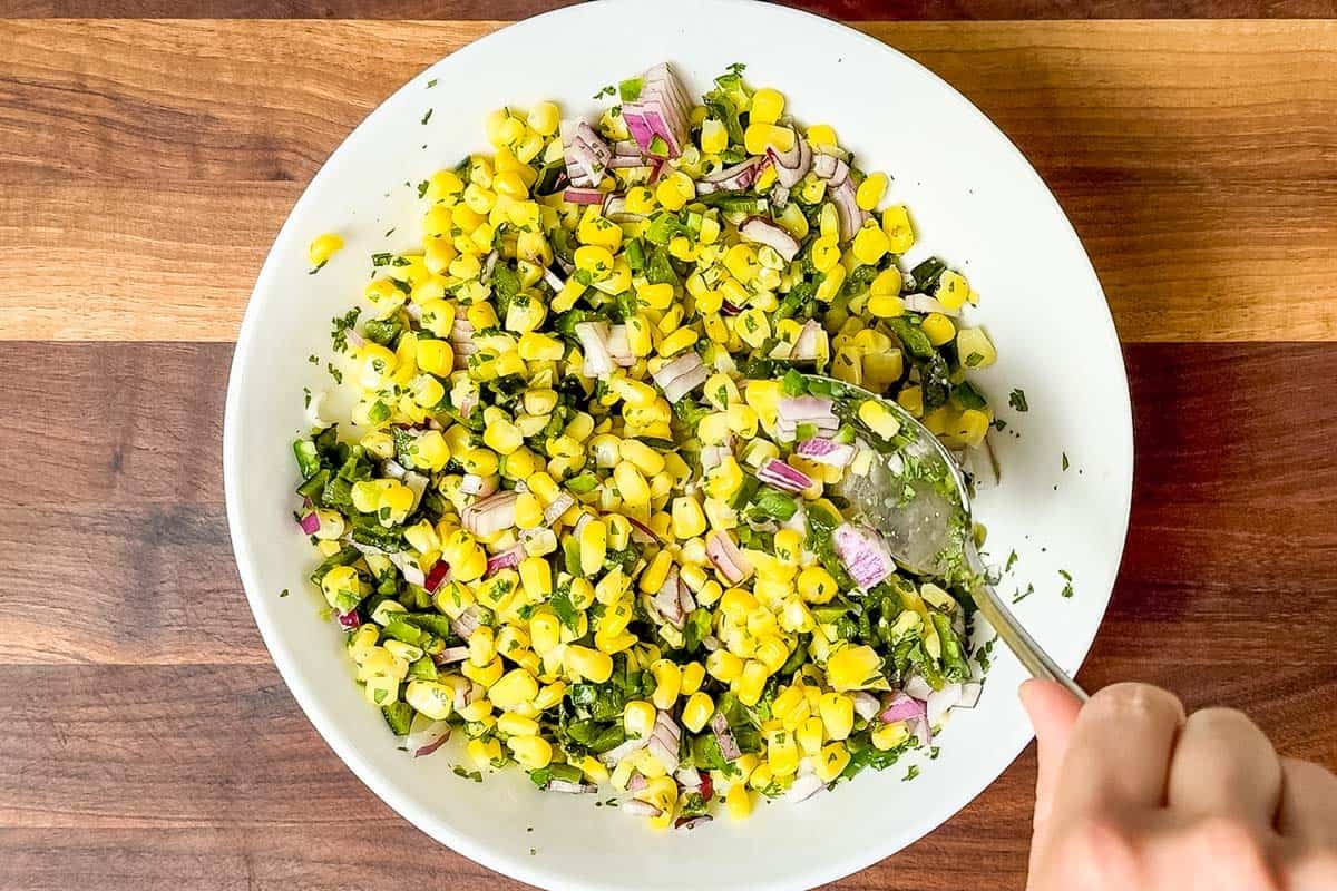 Mixing the corn salsa in a large white bowl with a metal spoon.