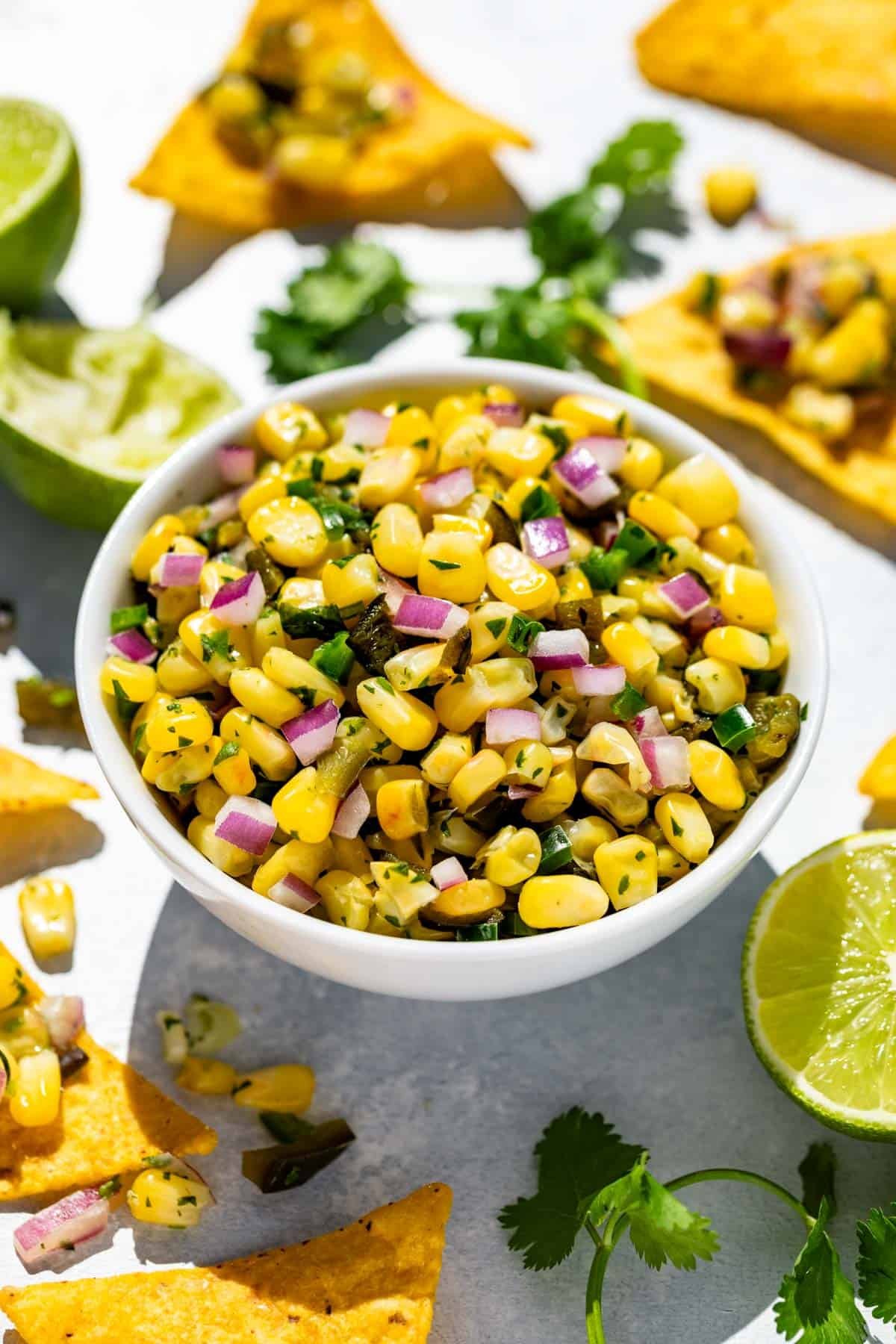 A white bowl filled with Roasted Chili Corn Salsa surrounded by corn chips and lime wedges.
