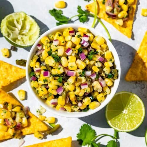 Finished Roasted Chili Corn Salsa in a white bowl surrounded by chips dipped into the salsa, lime wedges and cilantro sprigs.