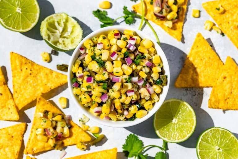 Finished Roasted Chili Corn Salsa in a white bowl surrounded by chips dipped into the salsa, lime wedges and cilantro sprigs.