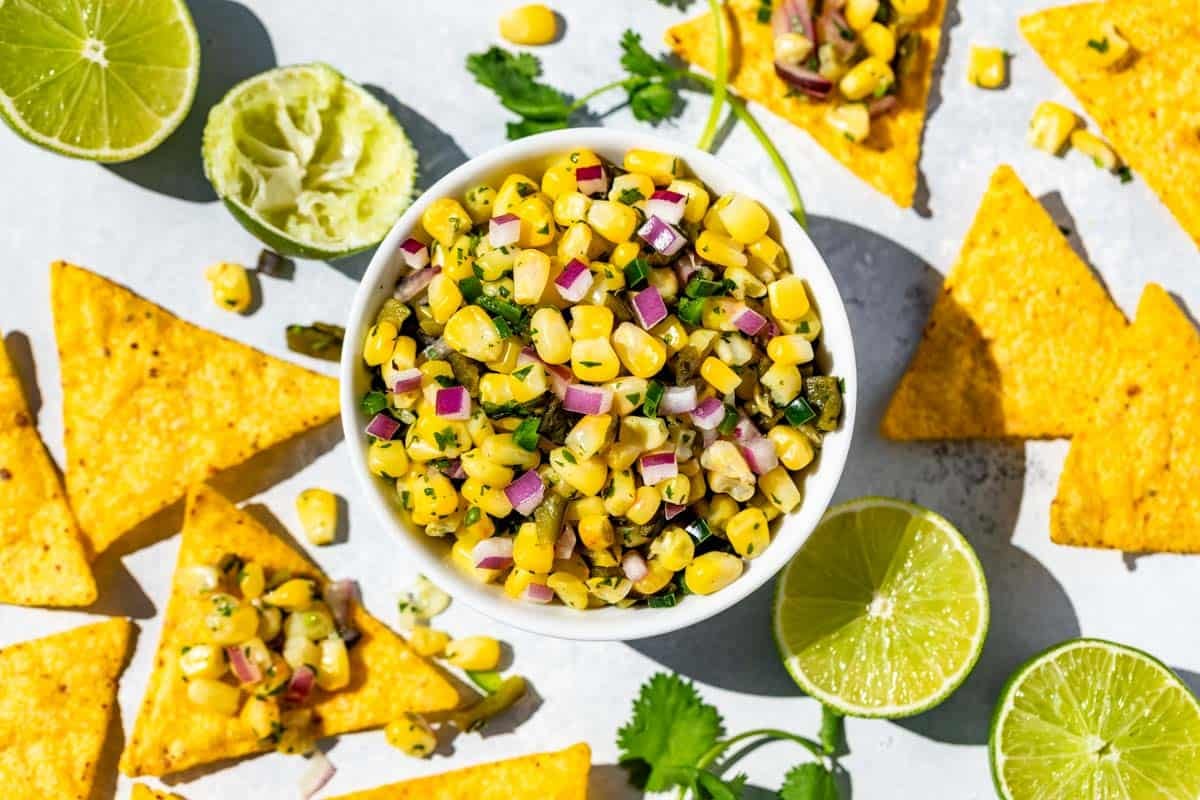 Finished Roasted Chili Corn Salsa in a white bowl surrounded by chips dipped into the salsa, lime wedges and cilantro sprigs.