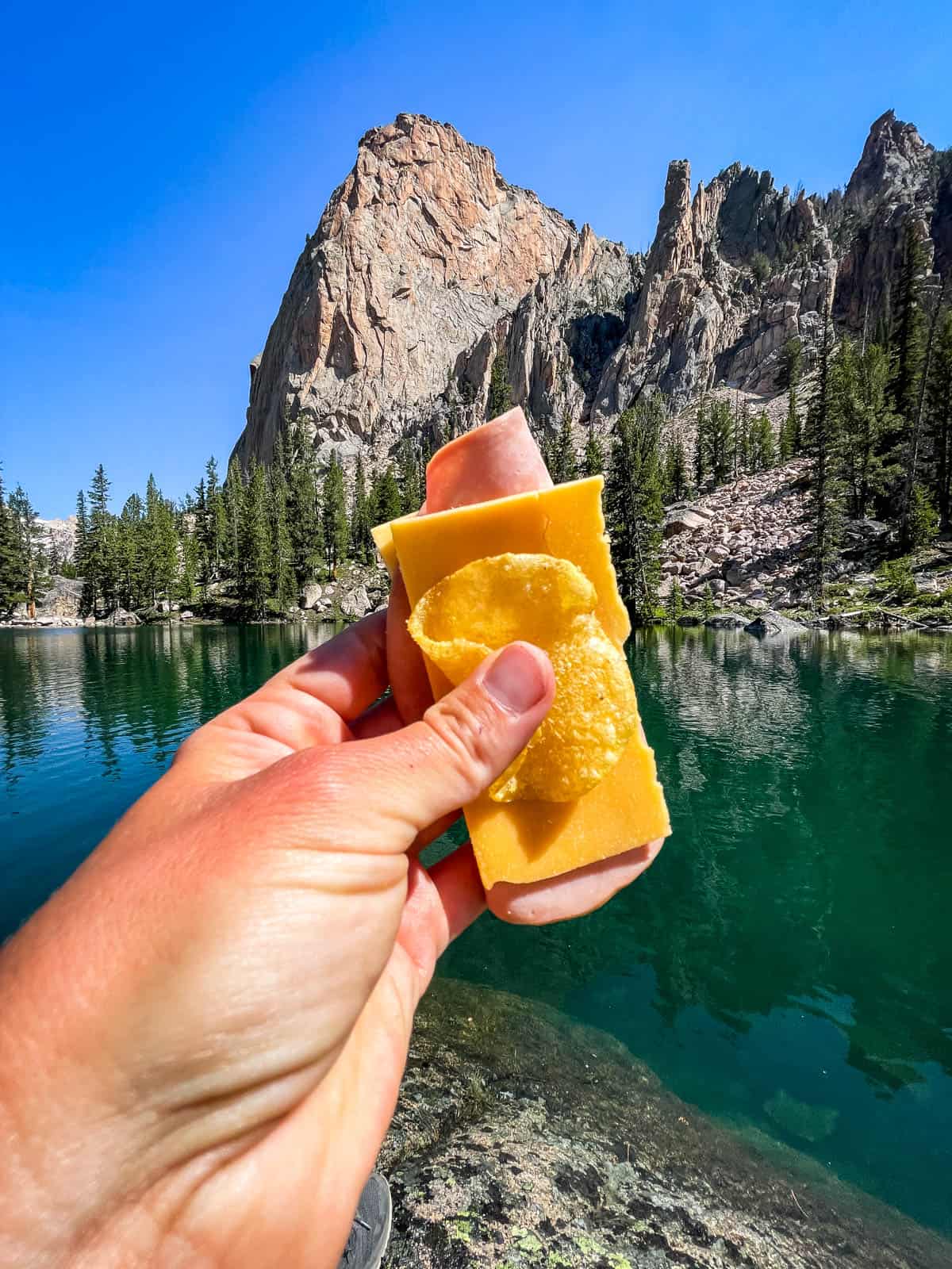 A hand holding sliced ham, cheddar, and potato chips at a remote backcountry mountain lake.