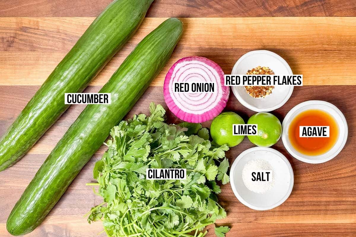 Cucumbers, cilantro, red onion, limes, red pepper flakes, sea salt, and agave nectar in bowls on a wood cutting board.