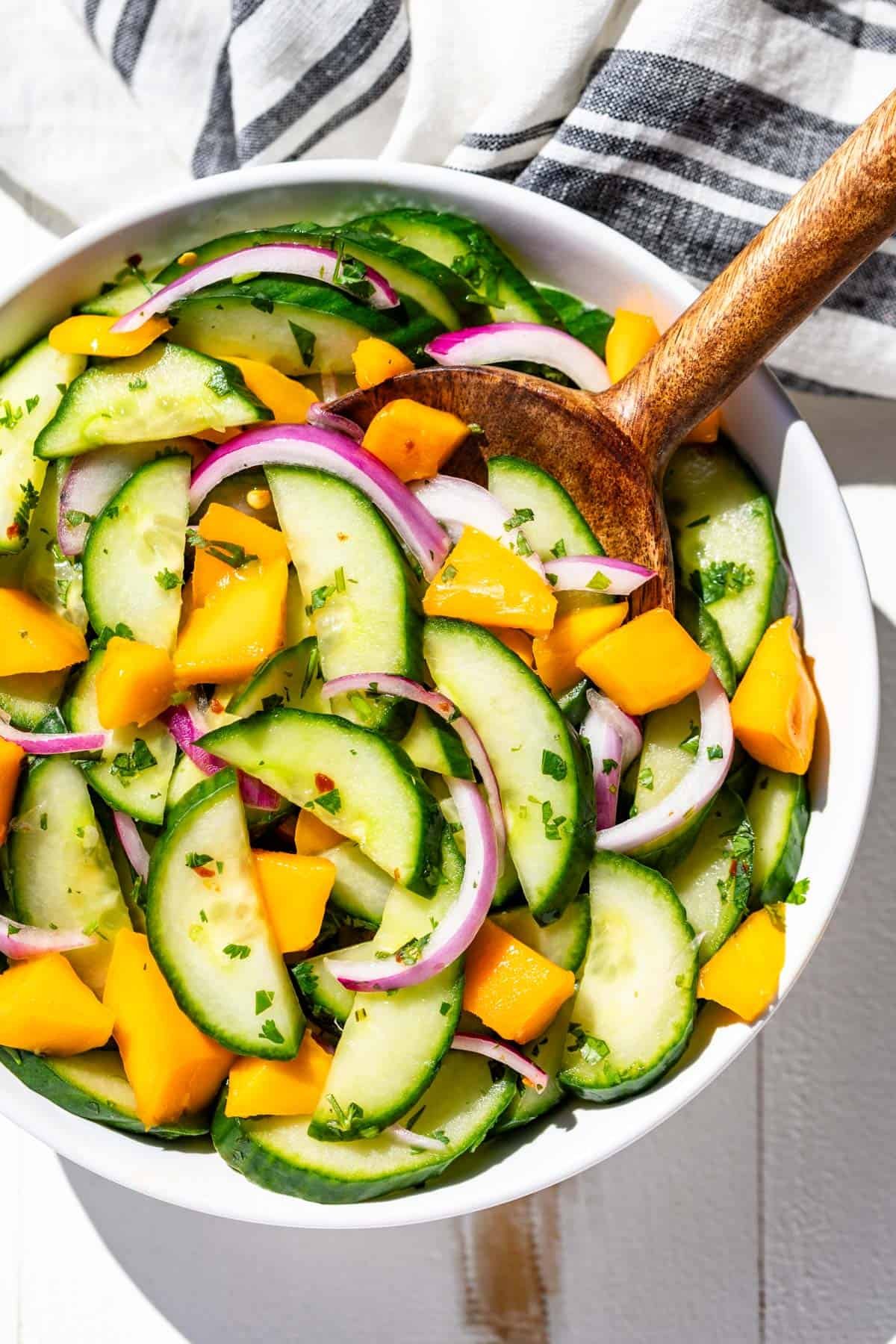 A white bowl filled with Asian cucumber salad with added mango chunks and a wood serving spoon in it.