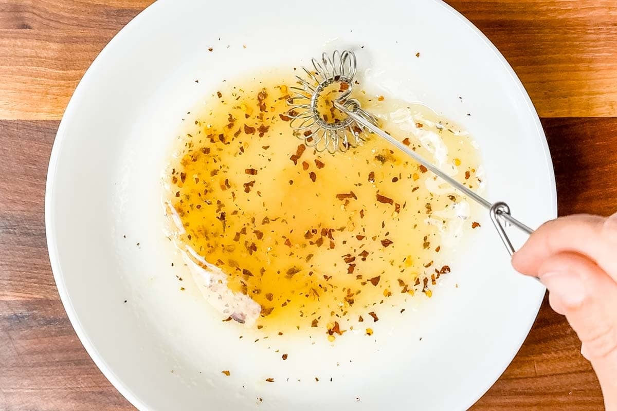 Whisking the salad dressing in a white bowl with a metal whisk on a wood cutting board.