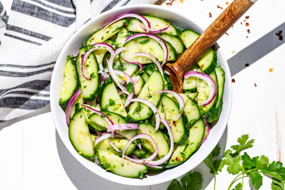 A white bowl filled with Asian cucumber salad with a wood serving spoon and topped with cilantro.