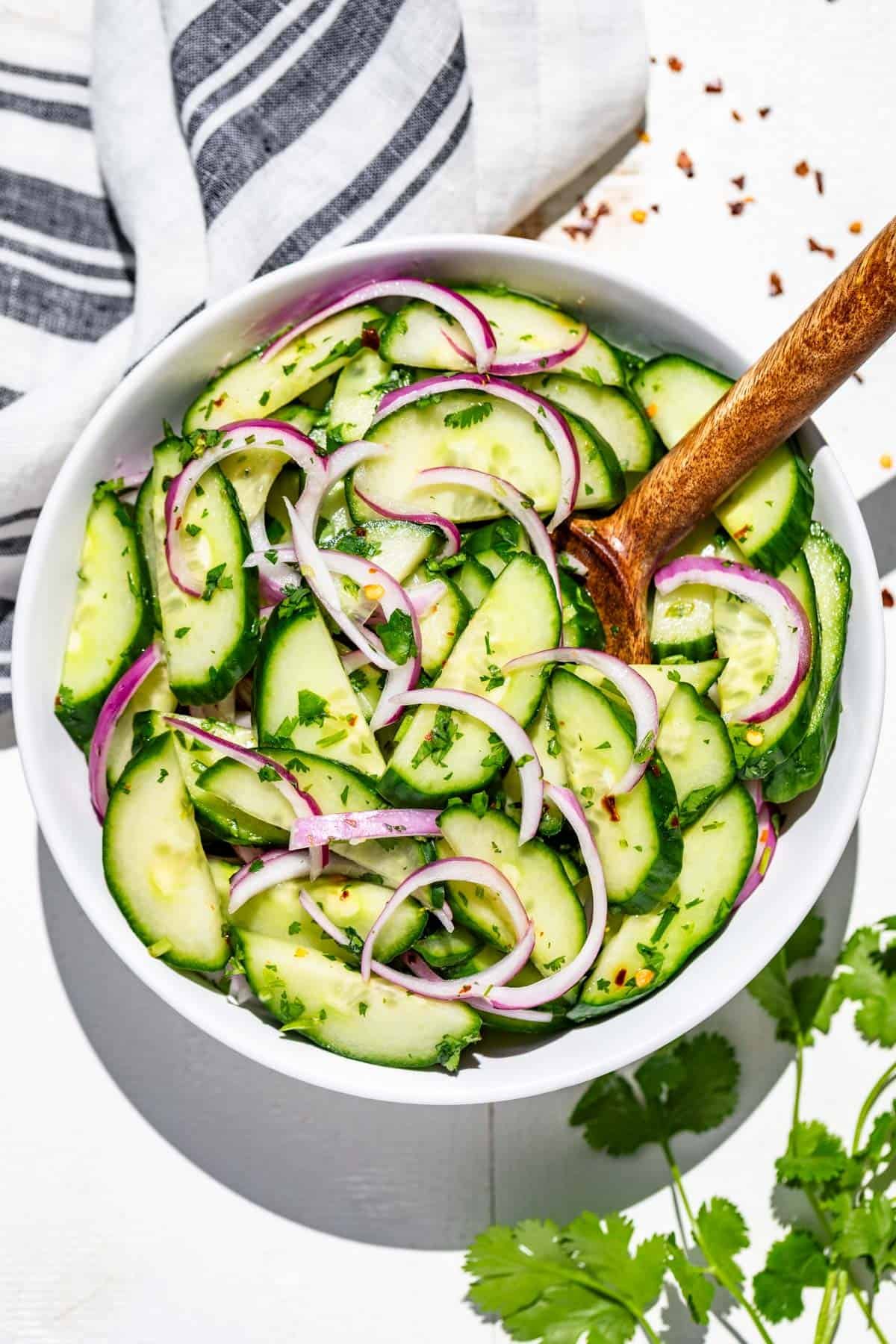 A white bowl filled with Asian cucumber salad topped with cilantro with a wood serving spoon in it.