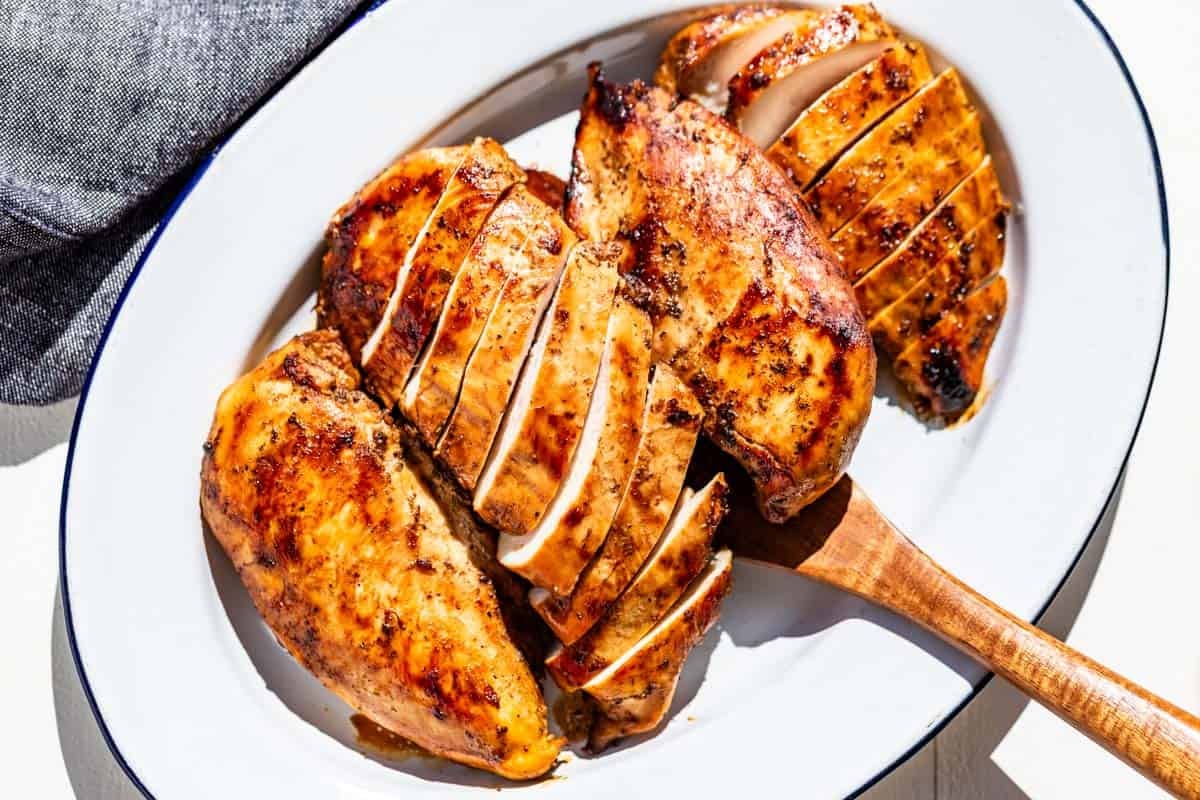 Four cooked chicken breasts on a blue rimmed white oval platter with a wood serving spatula.