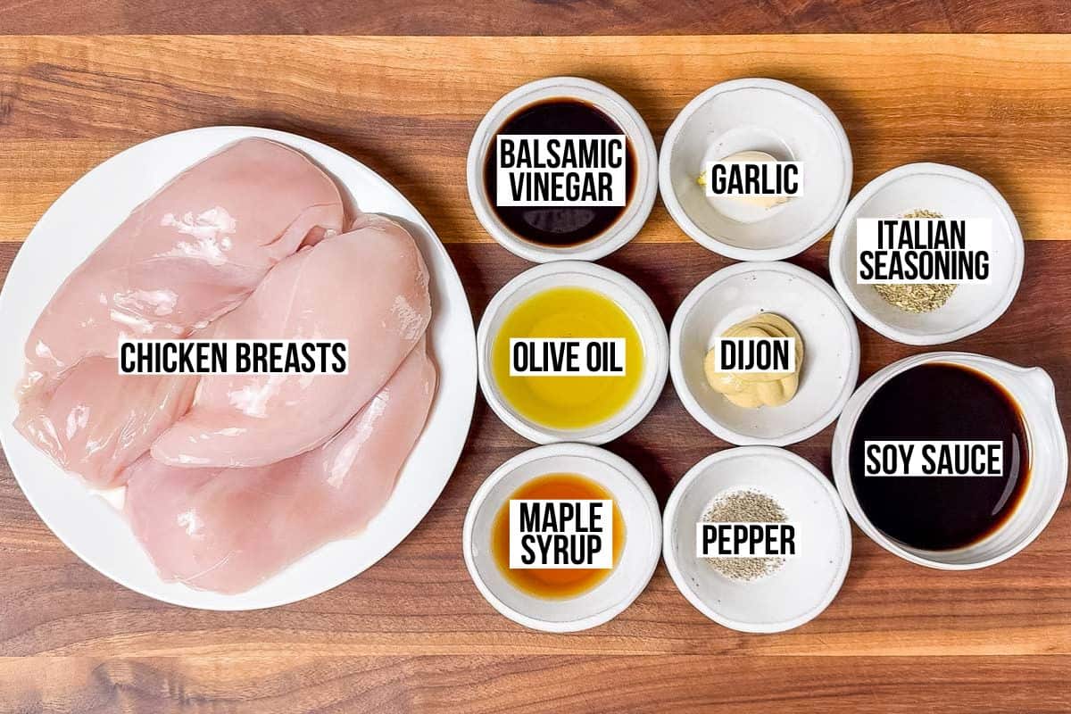 Chicken breasts, soy sauce, balsamic vinegar, maple syrup, Dijon mustard, garlic, Italian seasoning, and olive oil in bowls on a wood cutting board.
