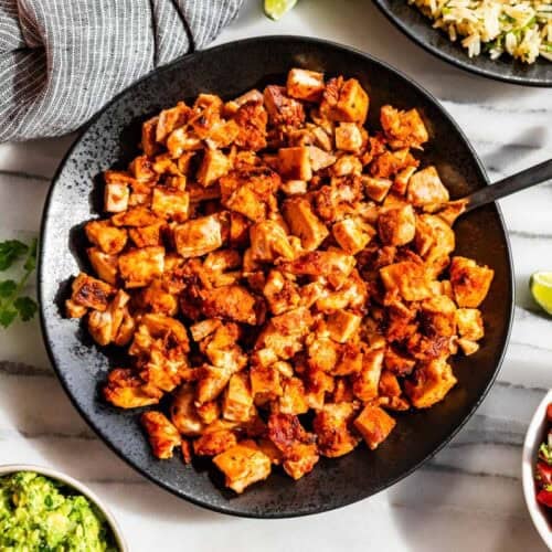 A black bowl of chopped Chipotle Chicken surrounded by bowls of Pico de Gallo, corn, guacamole, and rice with lime wedges.
