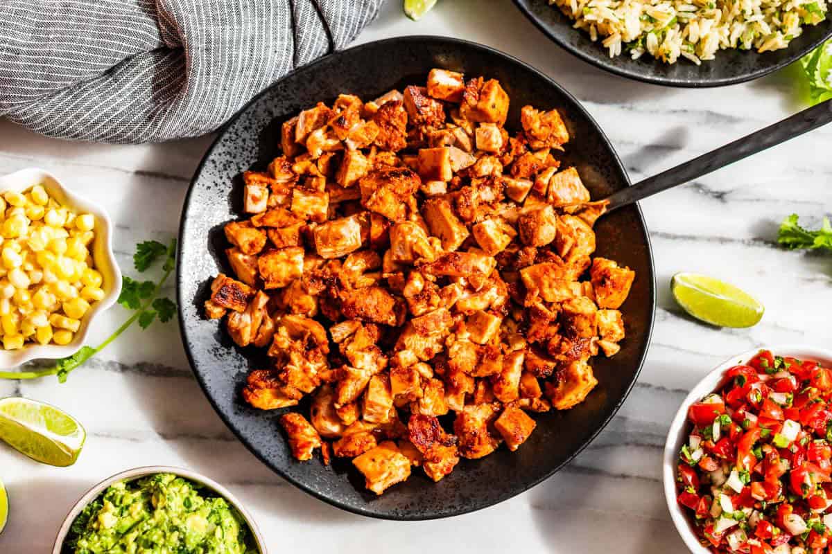 A black bowl of chopped Chipotle Chicken surrounded by bowls of Pico de Gallo, corn, guacamole, and rice with lime wedges.