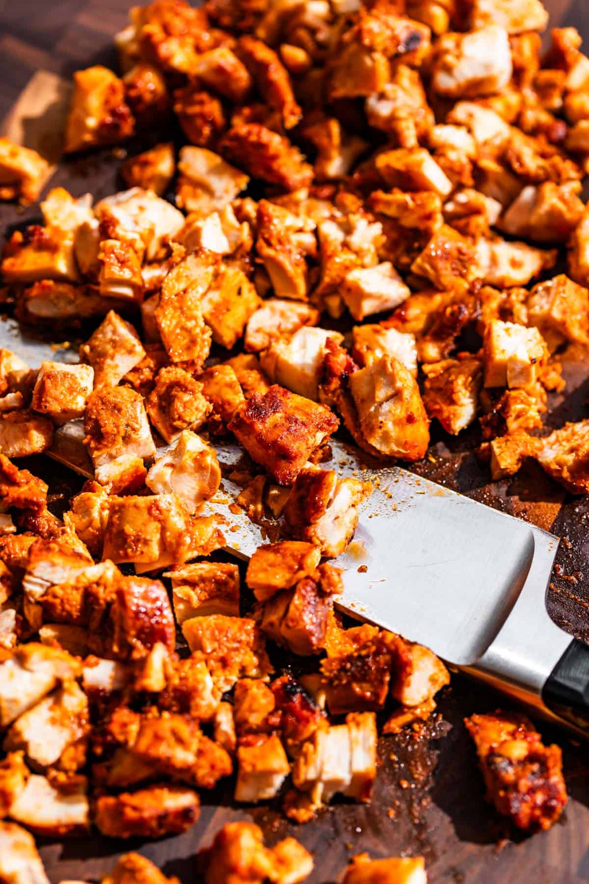A large chef's knife in the middle of chopped up Chipotle Chicken on a wood cutting board.