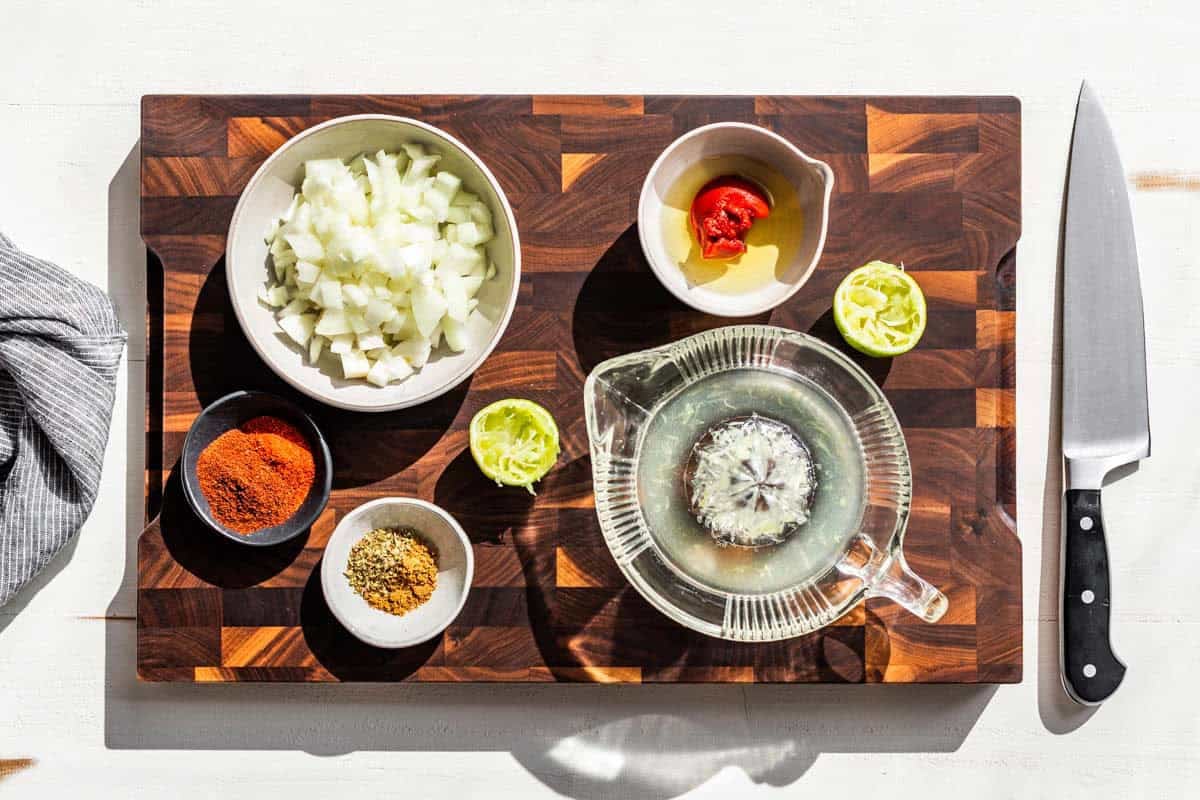 Chopped onions, lime juice, limes, cumin, tomato paste, and chipotle on a wood cutting board.