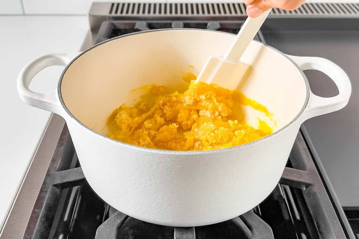 Mixing egg yolks and sugar with a spatula in a white pot on the stove top.