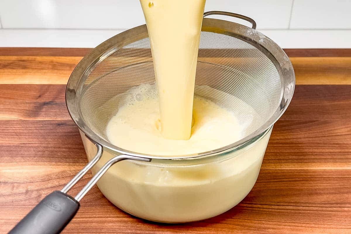 Pouring the finished custard through a fine mesh sieve into a glass bowl.