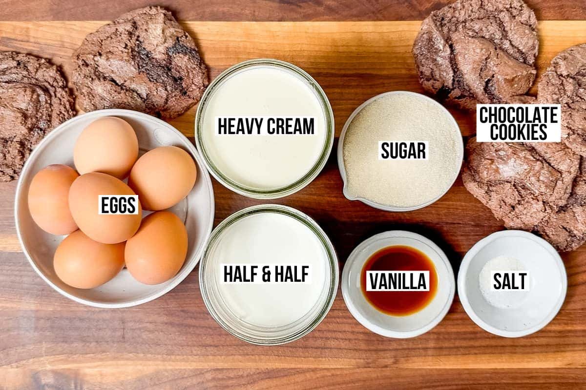Eggs, heavy cream, half and half, vanilla extract, salt, sugar, and chocolate cookies in containers on a wood cutting board.