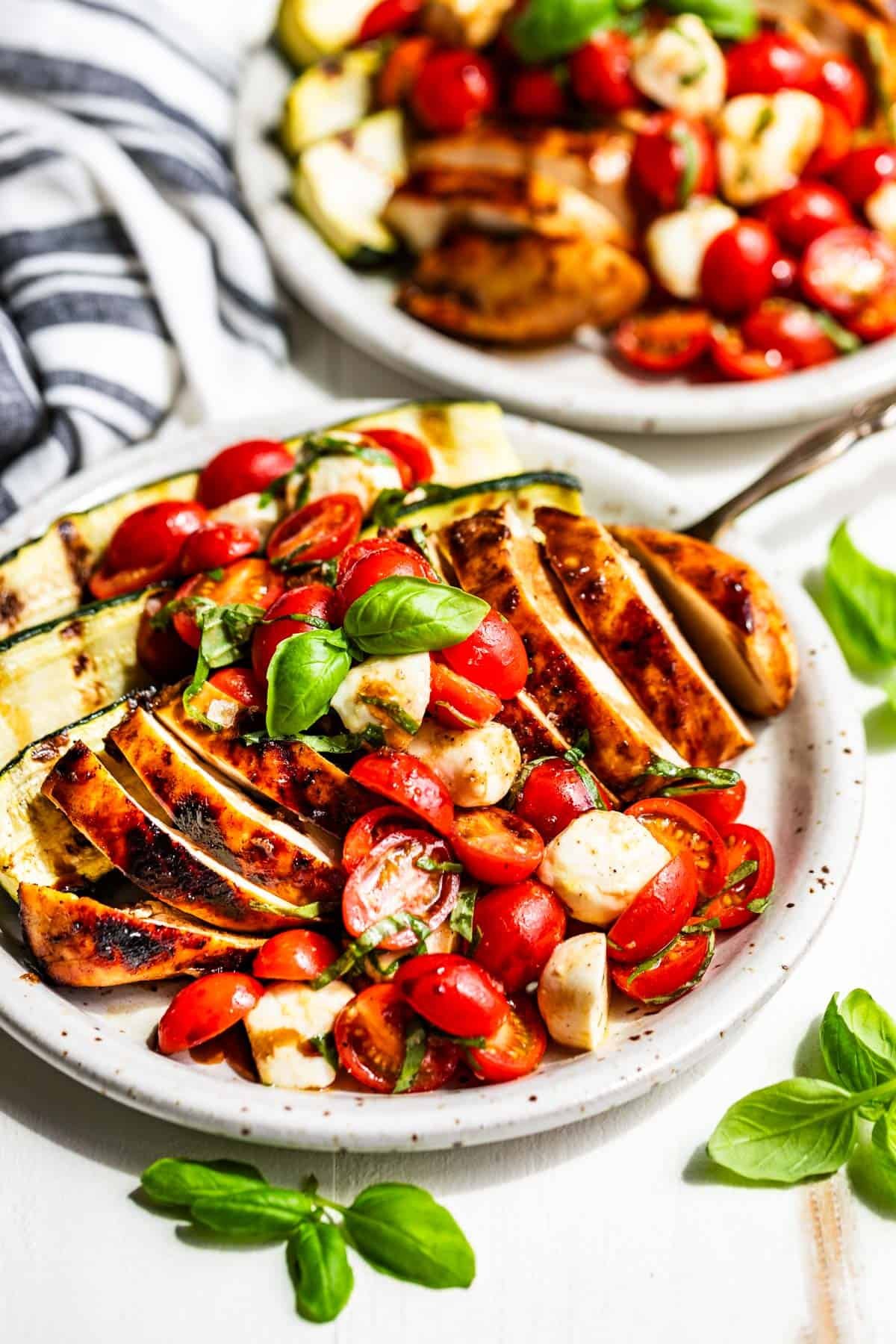 Two pottery plates of Balsamic Grilled Chicken topped with Caprese Salad with a blue and white striped linen on the side with basil sprigs around the plates.