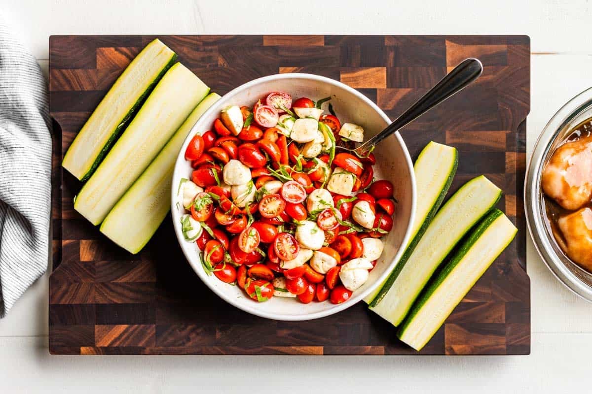 Caprese Salad in a mixing bowl with sliced zucchini on a wooden cutting board.