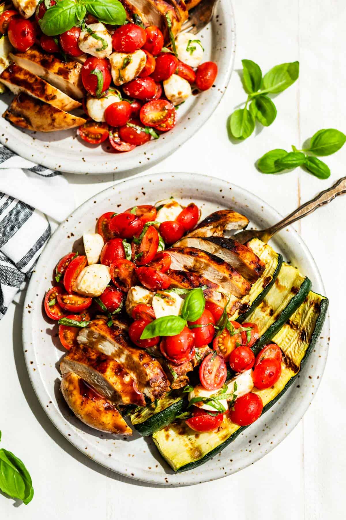 A straight down view of two plates of Balsamic Grilled Chicken topped with Caprese Salad on a white background with basil sprigs around it.