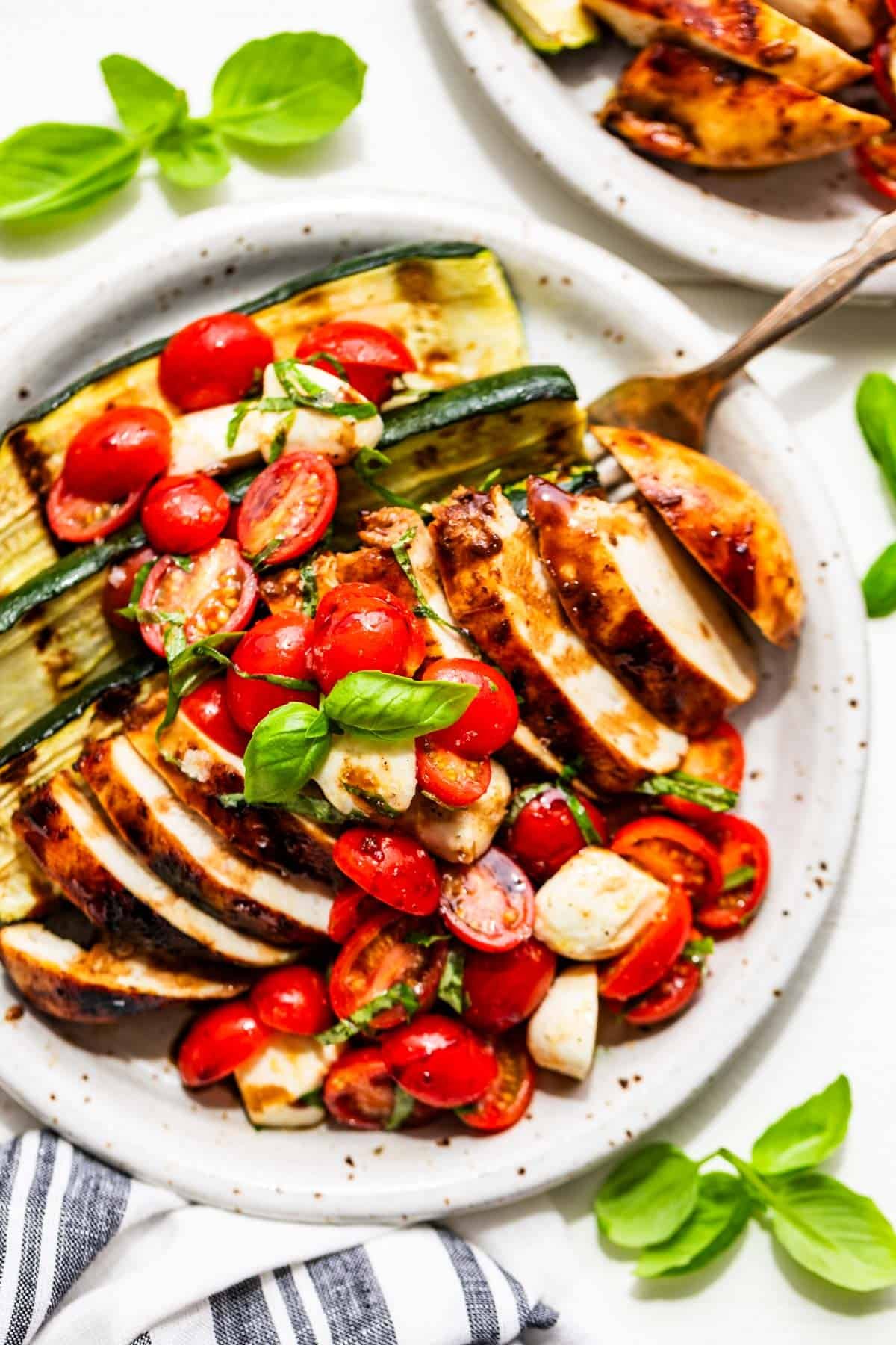 Balsamic Grilled Chicken topped with Caprese Salad on a pottery plate with basil sprigs around it and a blue and white striped linen next to the plate.