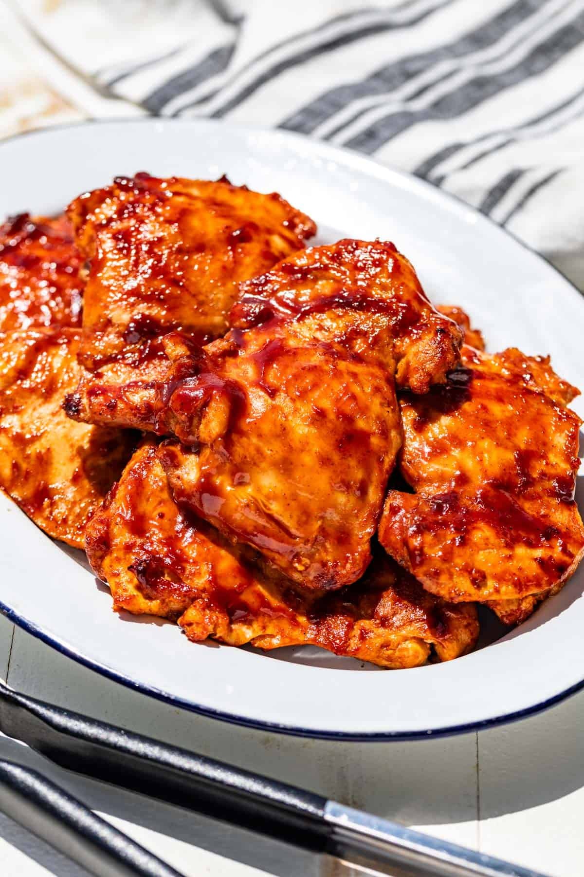 Grilled Chicken Thighs on a blue rimmed white oval platter with metal tongs in the front and a blue striped linen in the background.