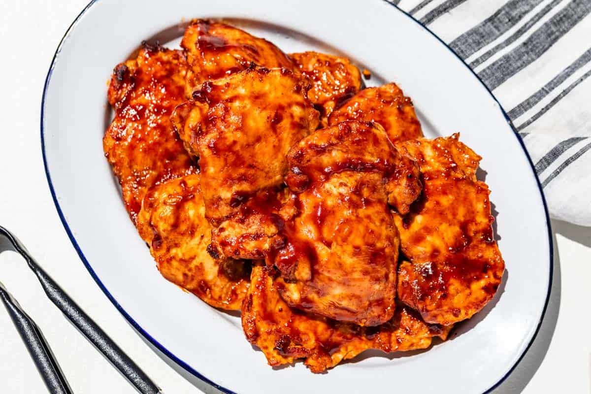 Finished grilled chicken thighs piled up on a blue rimmed white oval platter with metal tongs on the side.
