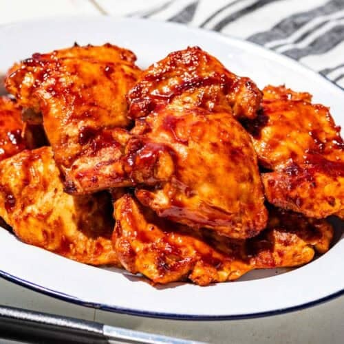 Grilled chicken thighs on a blue rimmed white platter with tongs in the front and a blue striped linen in the background.