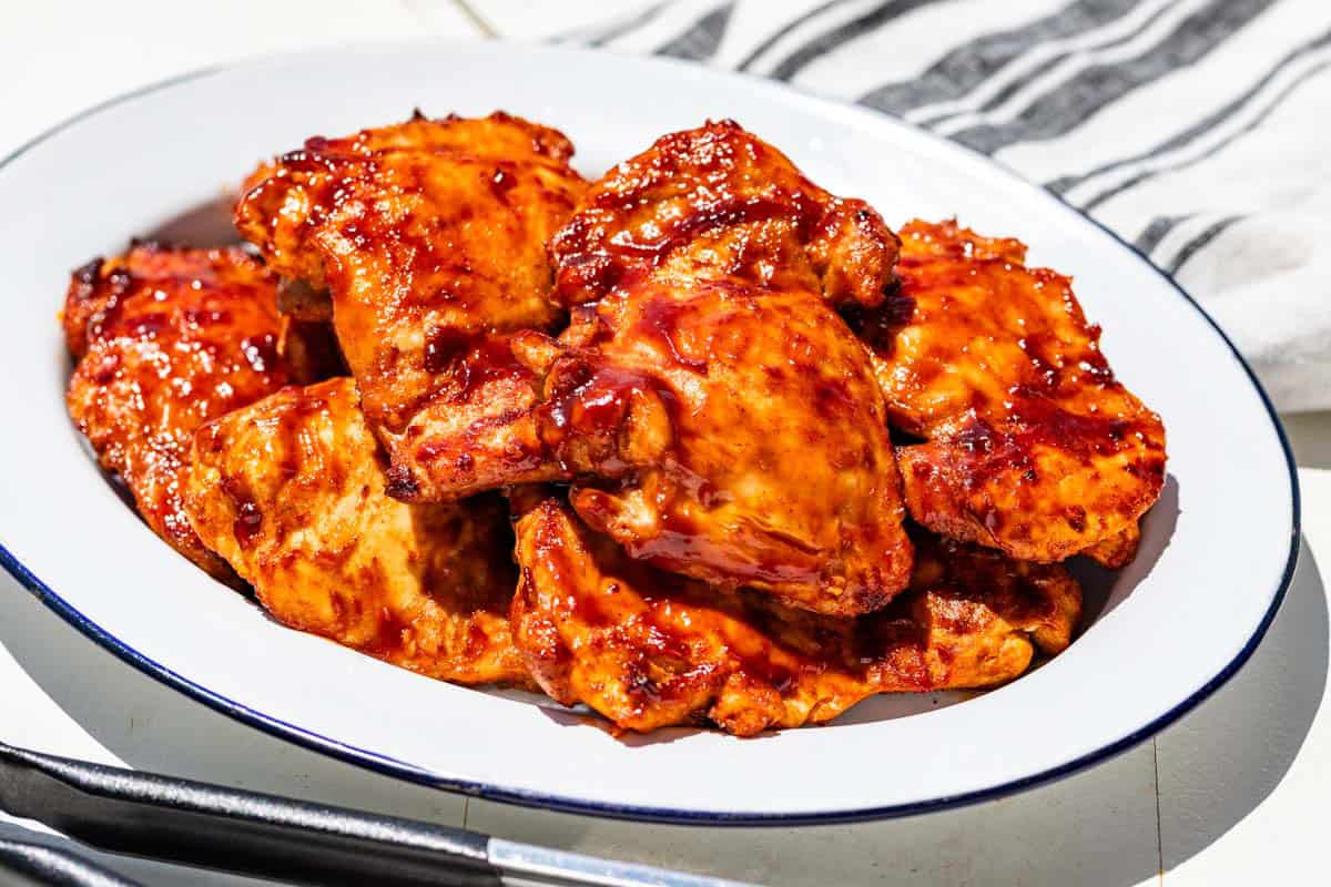 Grilled chicken thighs on a blue rimmed white platter with tongs in the front and a blue striped linen in the background.