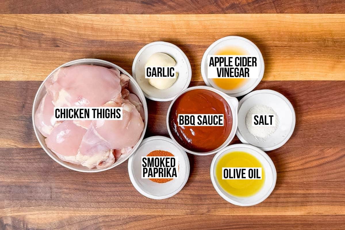 Chicken thighs, bbq sauce, smoked paprika, garlic, olive oil, apple cider vinegar, and salt in bowls on a wood cutting board.