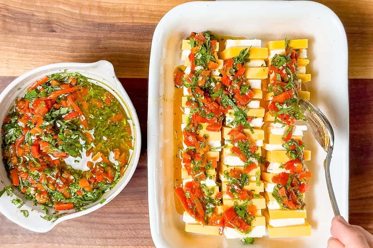 Spooning the red pepper herb marinade over the cheddar cheese and cream cheese squares in a baking dish.