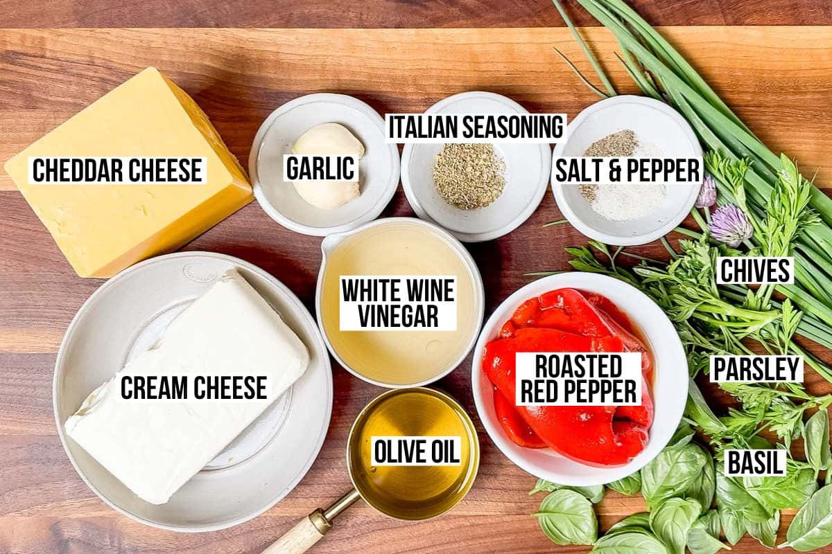 Cheddar cheese, cream cheese, garlic, roasted red bell pepper, white wine vinegar, Italian seasoning, basil, chives, and parsley in bowls on a wood cutting board.