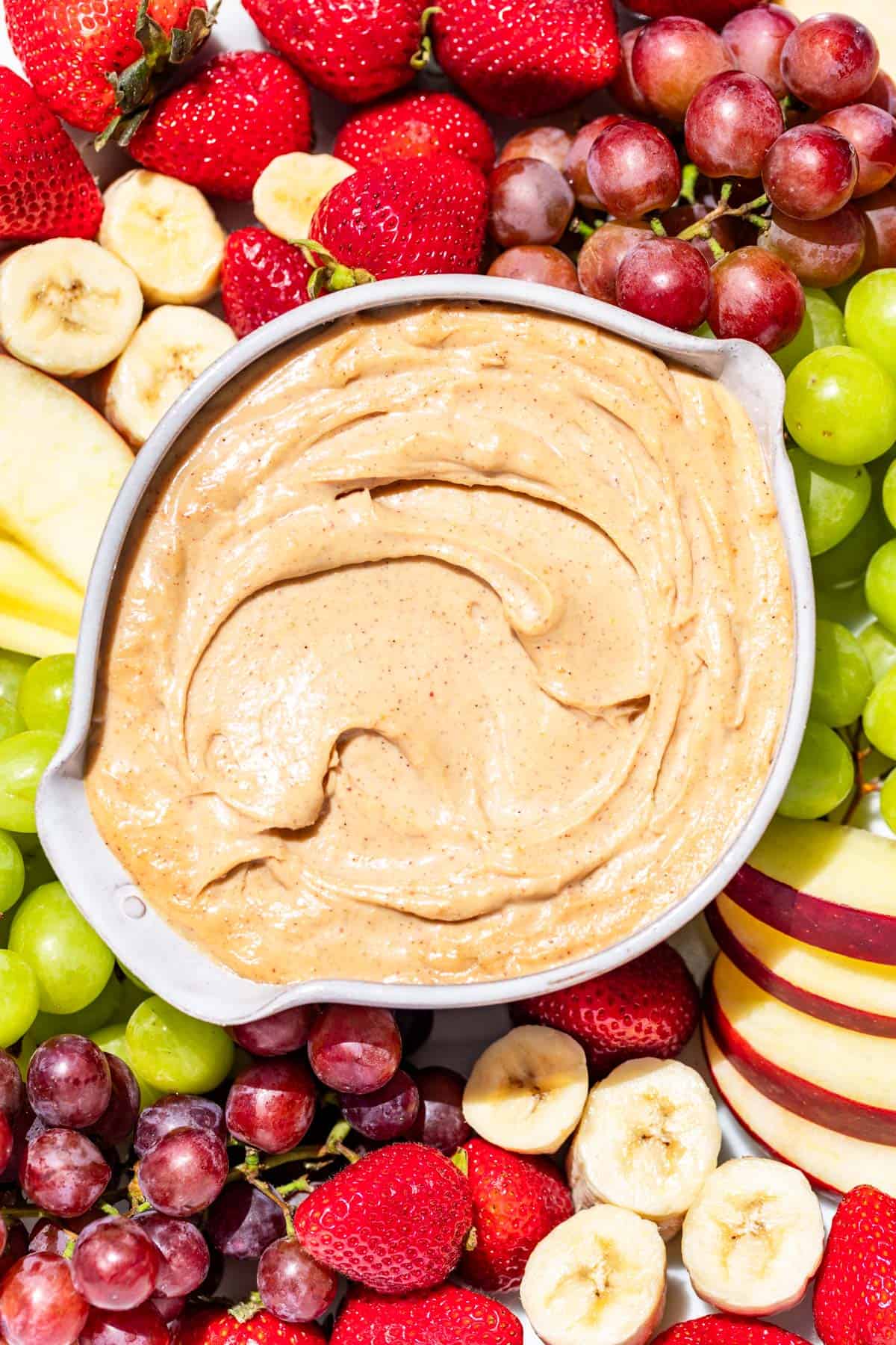 A pottery bowl of peanut butter dip surrounded by grapes, apple slices, strawberries, and banana slices.