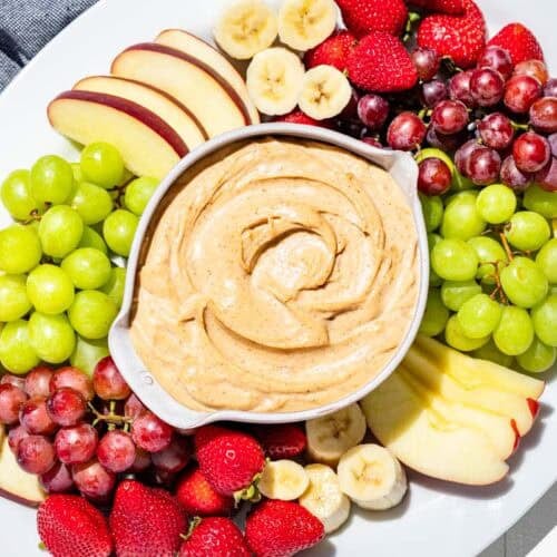 Finished peanut butter dip in a pottery bowl in the center of a large white platter surrounded by apple slices, grapes, strawberries, and banana slices.